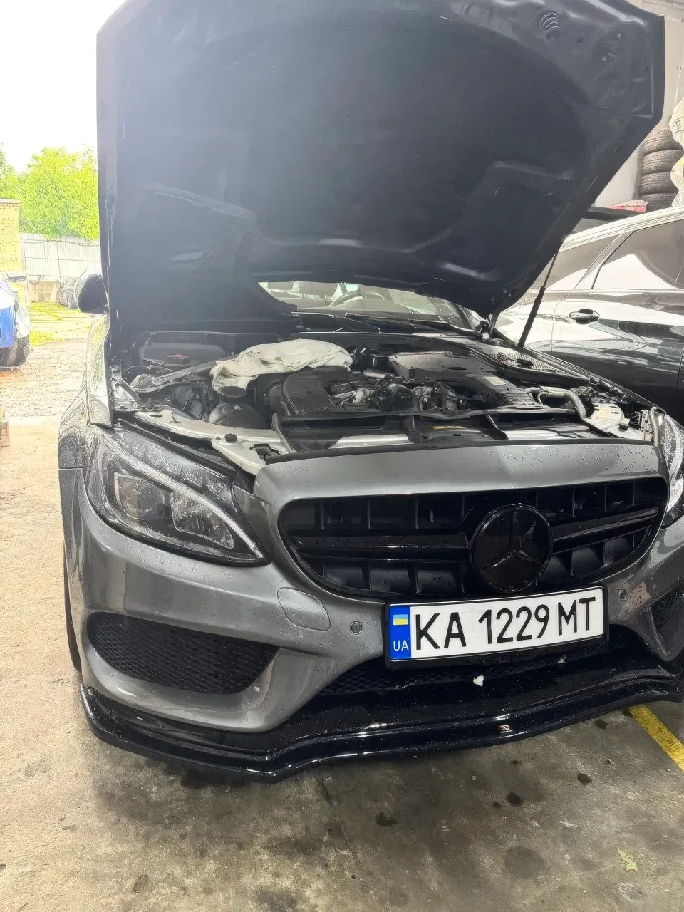 Спереду показано сірий Mercedes-Benz AMG із відкритим капотом і частково відкритим моторним відсіком. Людей у кадрі немає. Денне світло з входу змішується зі св