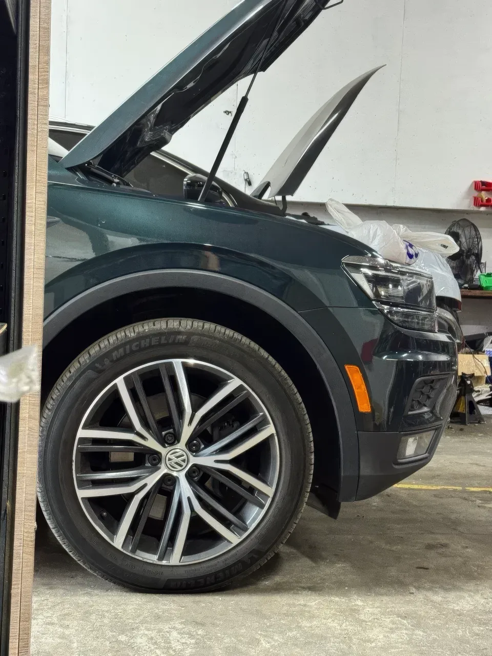 Великим планом показано переднє колесо та крило темно-зеленого Volkswagen Tiguan із відкритим капотом. Людей у кадрі немає. Штучне освітлення добре передає факт