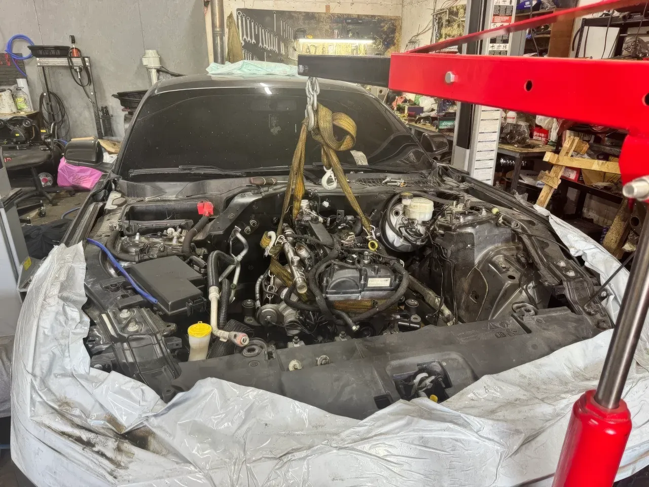 У майстерні показано моторний відсік Mercedes-Benz у процесі складного ремонту, над двигуном проходять стропи від червоного гідравлічного крана. Людей у кадрі н